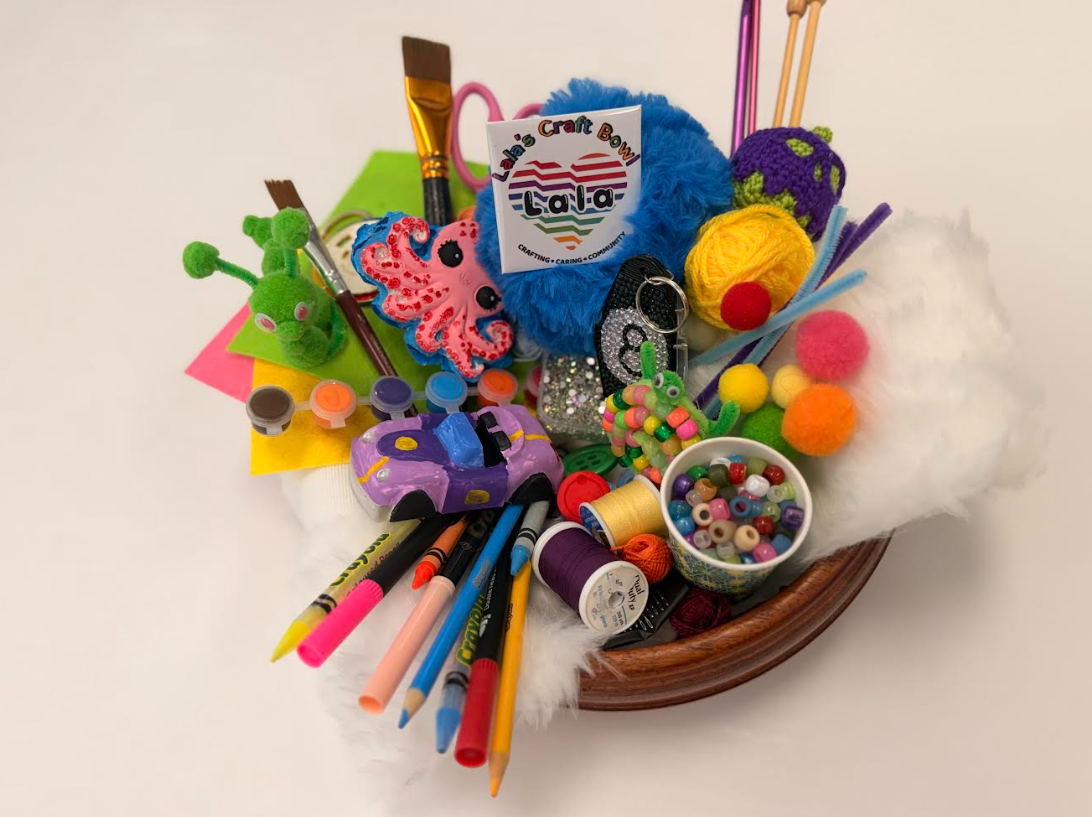 A wooden bowl overflowing with colourful craft supplies — yarn, beads, crayons, markers, paintbrushes, pom-poms, and handmade creatures.